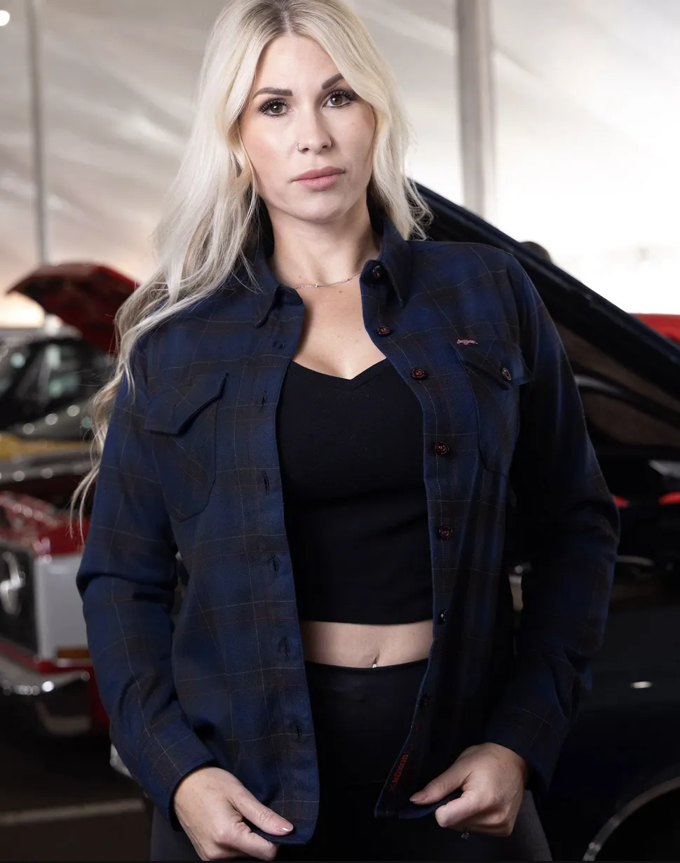 A woman with long blonde hair wearing an open dark blue and charcoal plaid Dixxon flannel with subtle red pinstripes over a black cropped tank top. She is standing in front of a classic car with the hood popped.