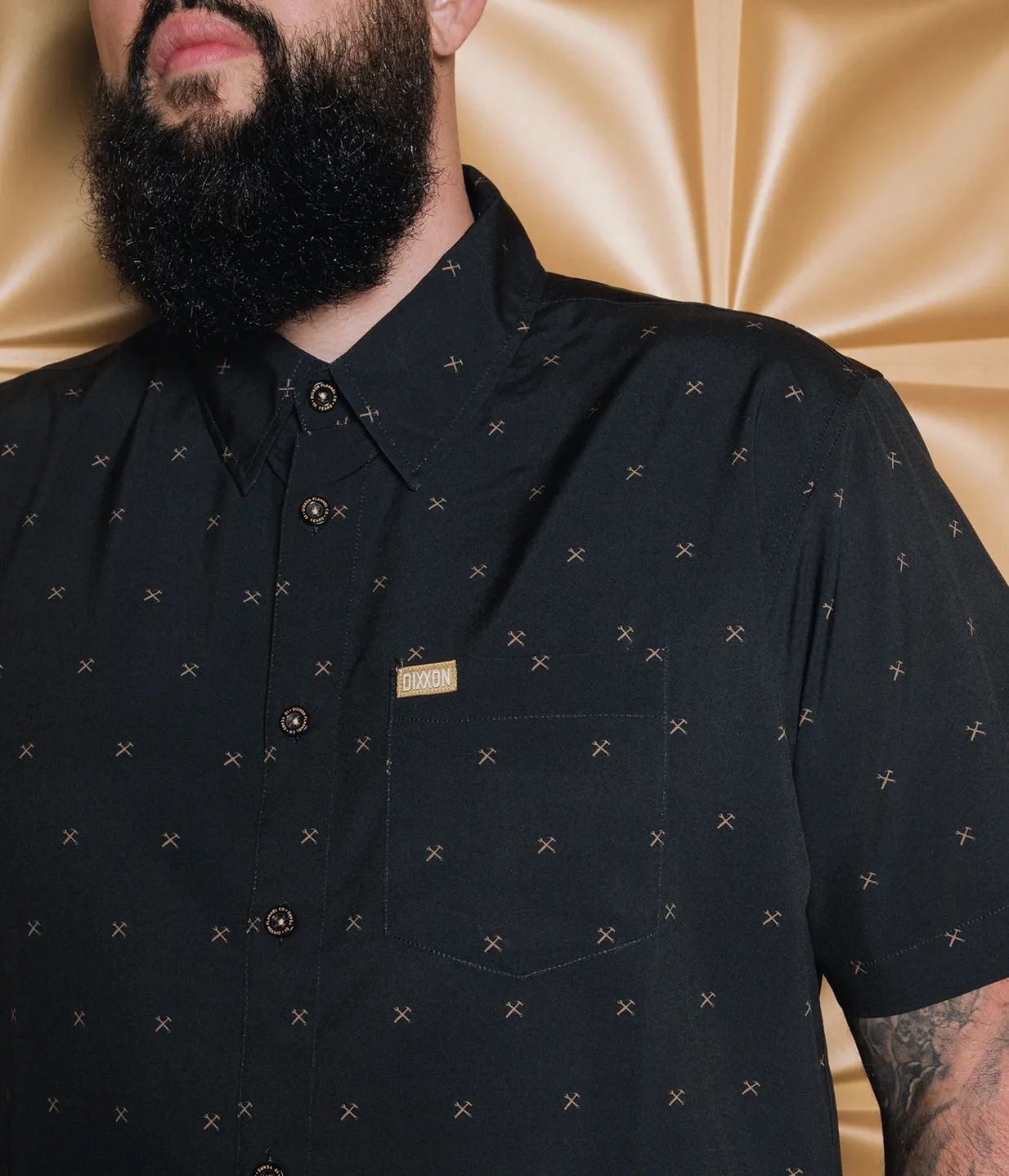 A close-up of a man with a large beard wearing a black short-sleeved Dixxon party shirt. The shirt is covered in a small, repeating gold "Work Hard" logo consisting of a crossed wrench and hammer. The shirt features a gold Dixxon logo tag on the chest pocket and custom Dixxon buttons.