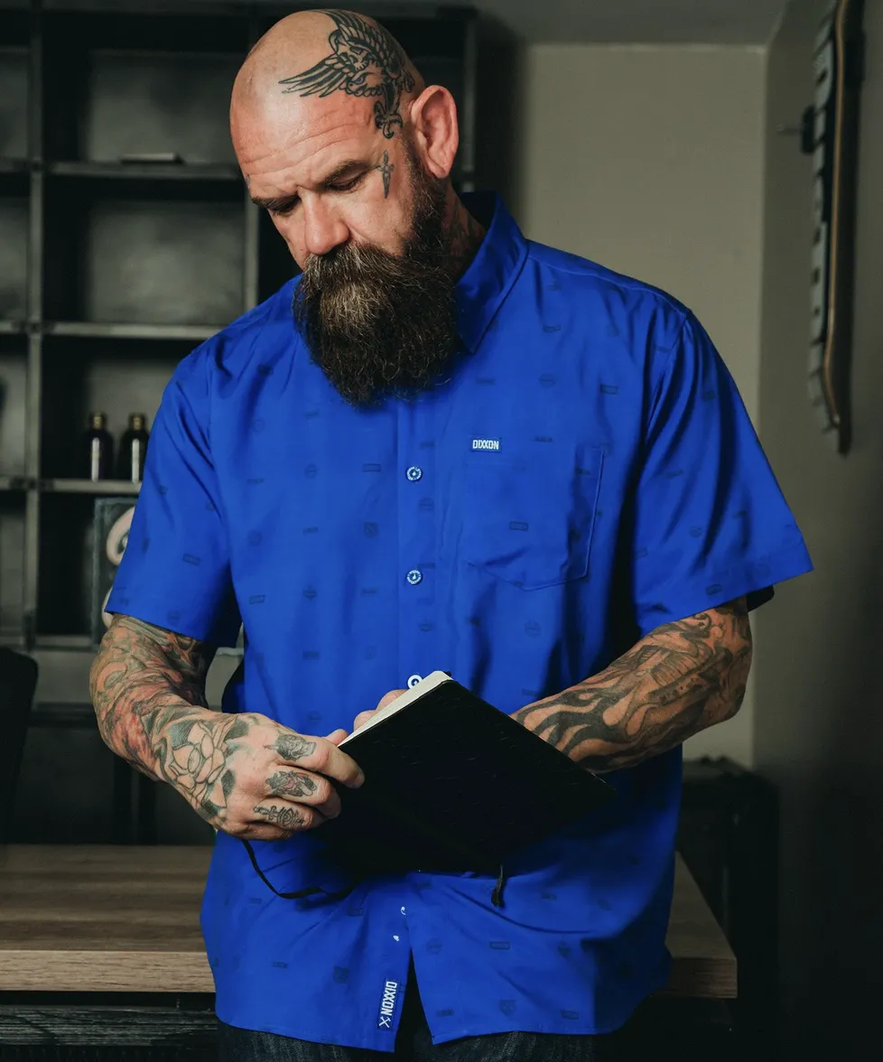 A man with a tattooed head and beard wears a royal blue Dixxon party shirt with a subtle, all-over repeating logo print. He is focused on writing in a black notebook in a workshop setting.