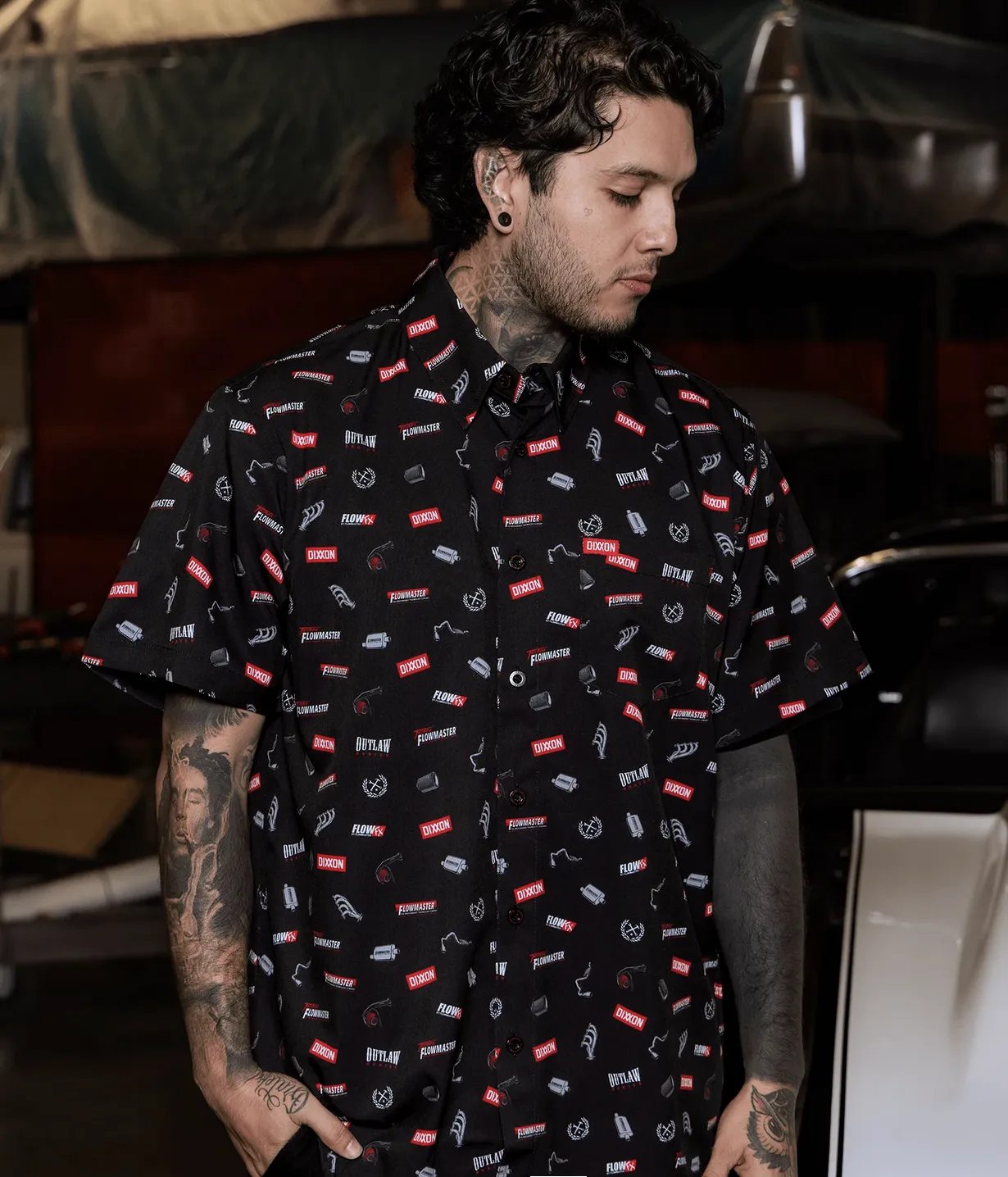 A man with visible neck and arm tattoos wearing a black short-sleeve Dixxon party shirt featuring a repeating "all-over print" pattern of red and white racing-inspired logos, including "Flowmaster" and "Outlaw". The setting appears to be a custom car workshop.