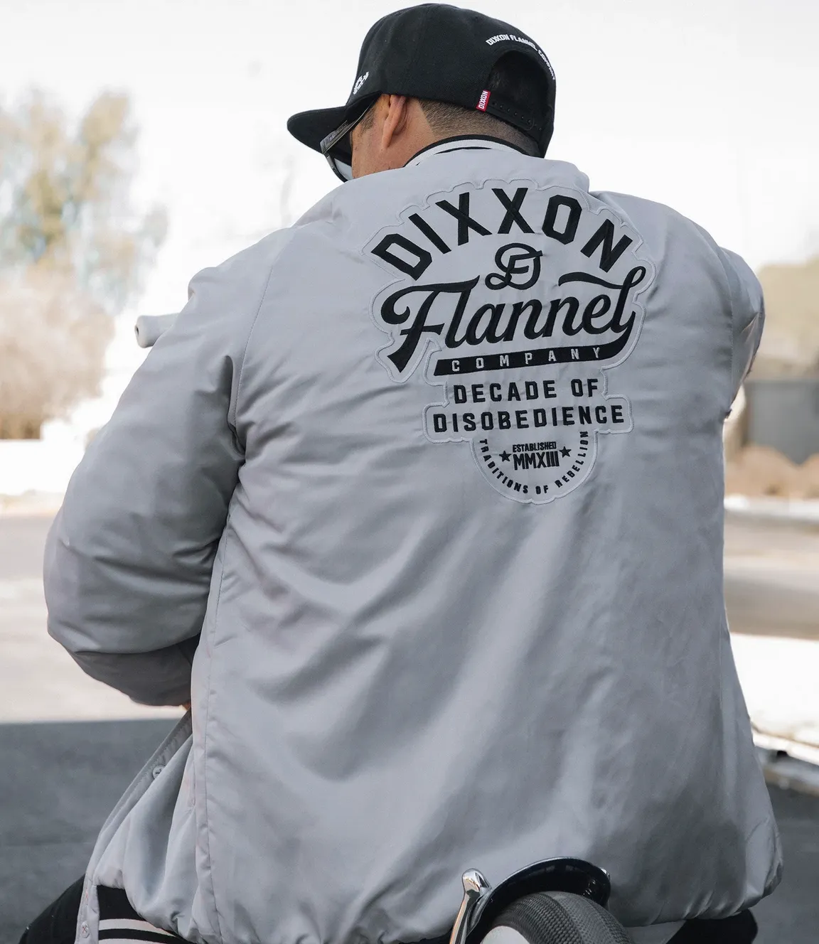 Rear view of a person sitting on a motorcycle wearing a light gray Varsity Jacket featuring large embroidered black text that reads "DIXXON Flannel Company DECADE OF DISOBEDIENCE