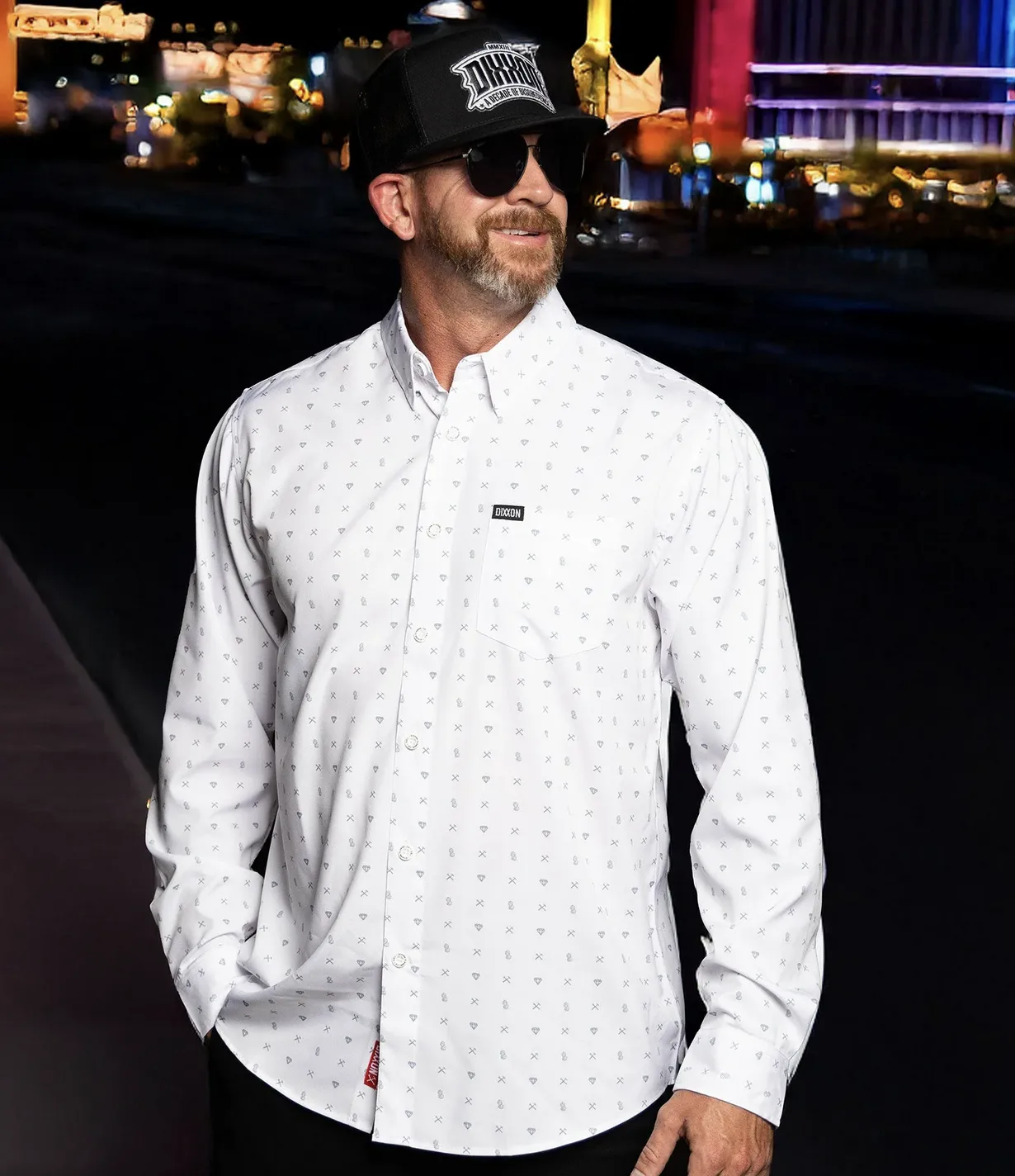 A man at night wearing a white long-sleeved button-down shirt from the Dixxon Office collection. The shirt features a subtle, all-over diamond and "Work Hard/Stay Humble" logo pattern. He is also wearing a black Dixxon "A Decade of Defiance" hat and dark sunglasses.