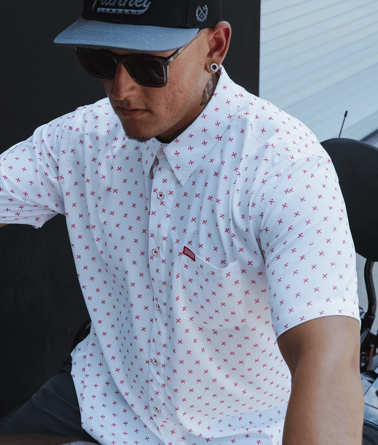 A man wearing a white Dixxon short-sleeve party shirt featuring an all-over red "X" pattern. He is wearing a black and grey Dixxon snapback hat and dark sunglasses. The shirt clearly shows the red Dixxon brand tag on the left chest pocket.