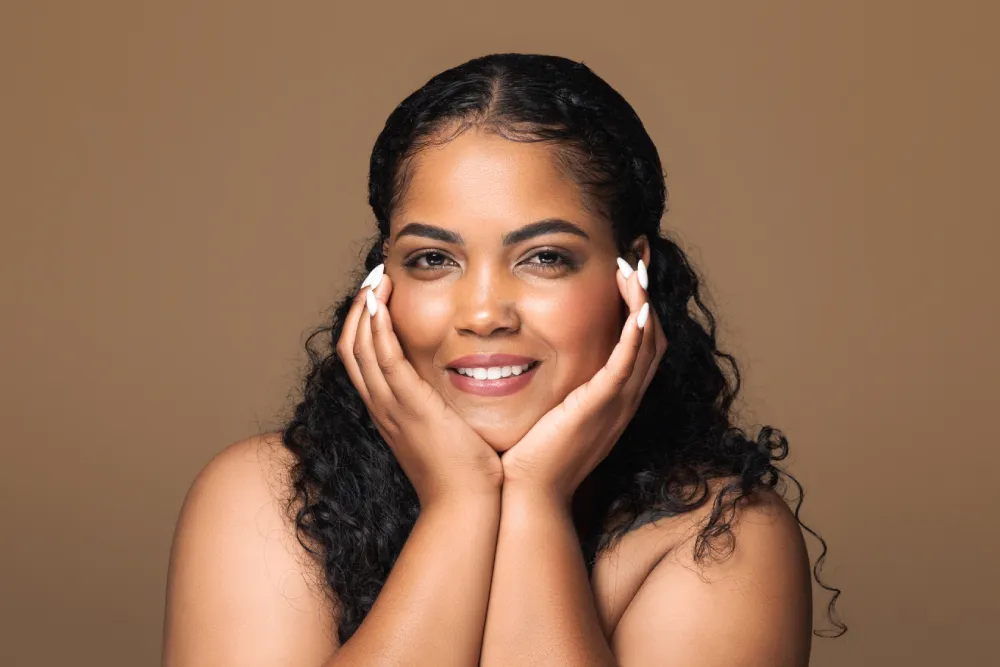 A beautiful woman with dark, curly hair and a warm smile gently cups her face with both hands, looking content and well-cared for. Her radiant expression highlights the importance of self-care, a vital topic for any new moms. The image is set against a soft, tan background, emphasizing her happy and glowing look.