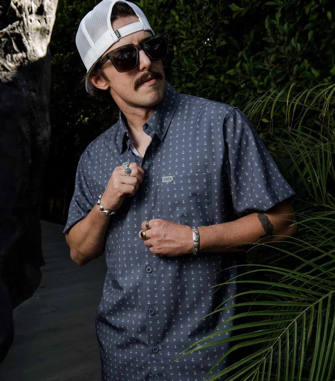 A close-up shot of a man with a mustache wearing a grey button-down shirt with a clean, repeating white anchor and cross print. He is adjusting his shirt near palm leaves, wearing sunglasses and a white Dixxon mesh-back cap.
