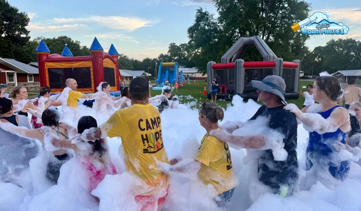A large group of children and adults participating in a foam event at an outdoor camp. They are standing in a thick layer of white foam, forming a line or circle with their hands on each other's shoulders. In the background, there are various inflatable attractions, including a colorful bounce house and a slide, under a twilight sky. A logo for "hilltop foam parties" is visible in the upper right corner.