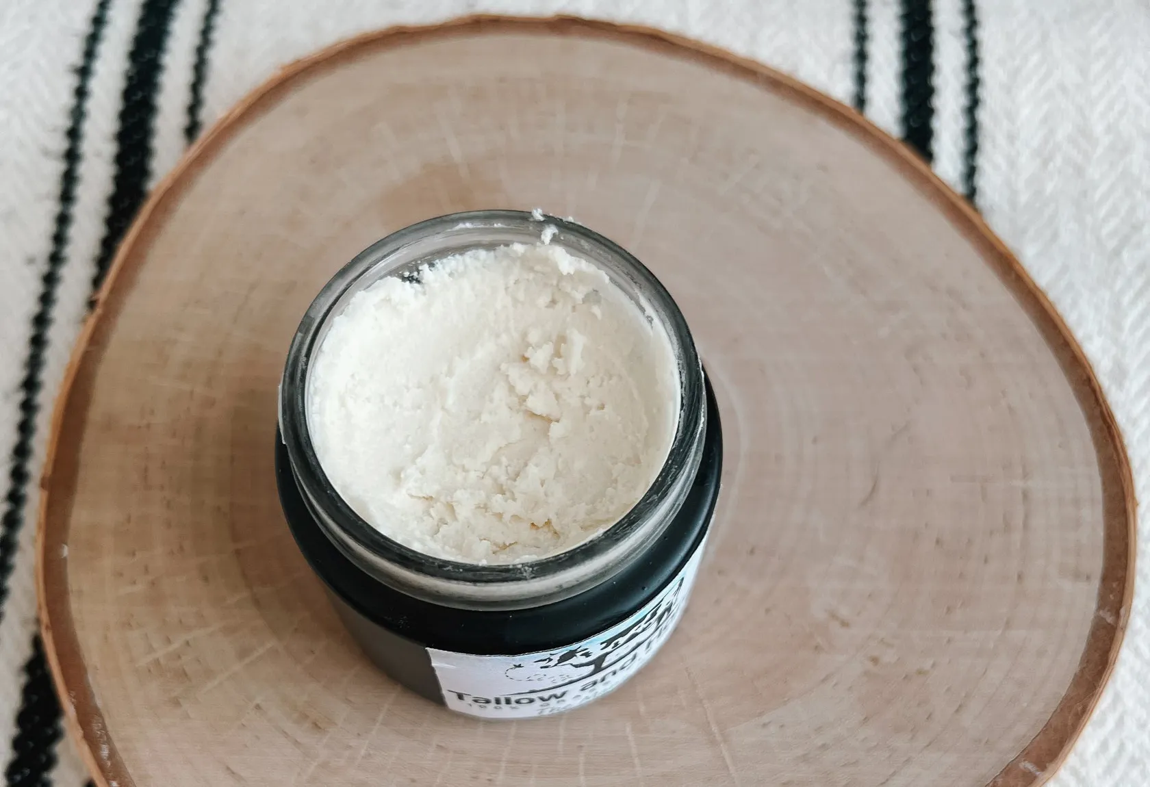 An open jar of whipped white Tallow Products resting on a natural wood slice with a rustic striped background.