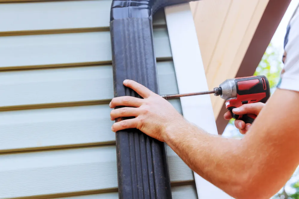 A close-up view of a worker using a power drill to secure a black metal downspout to the side of a house with light green siding during the installation of a gutter system.