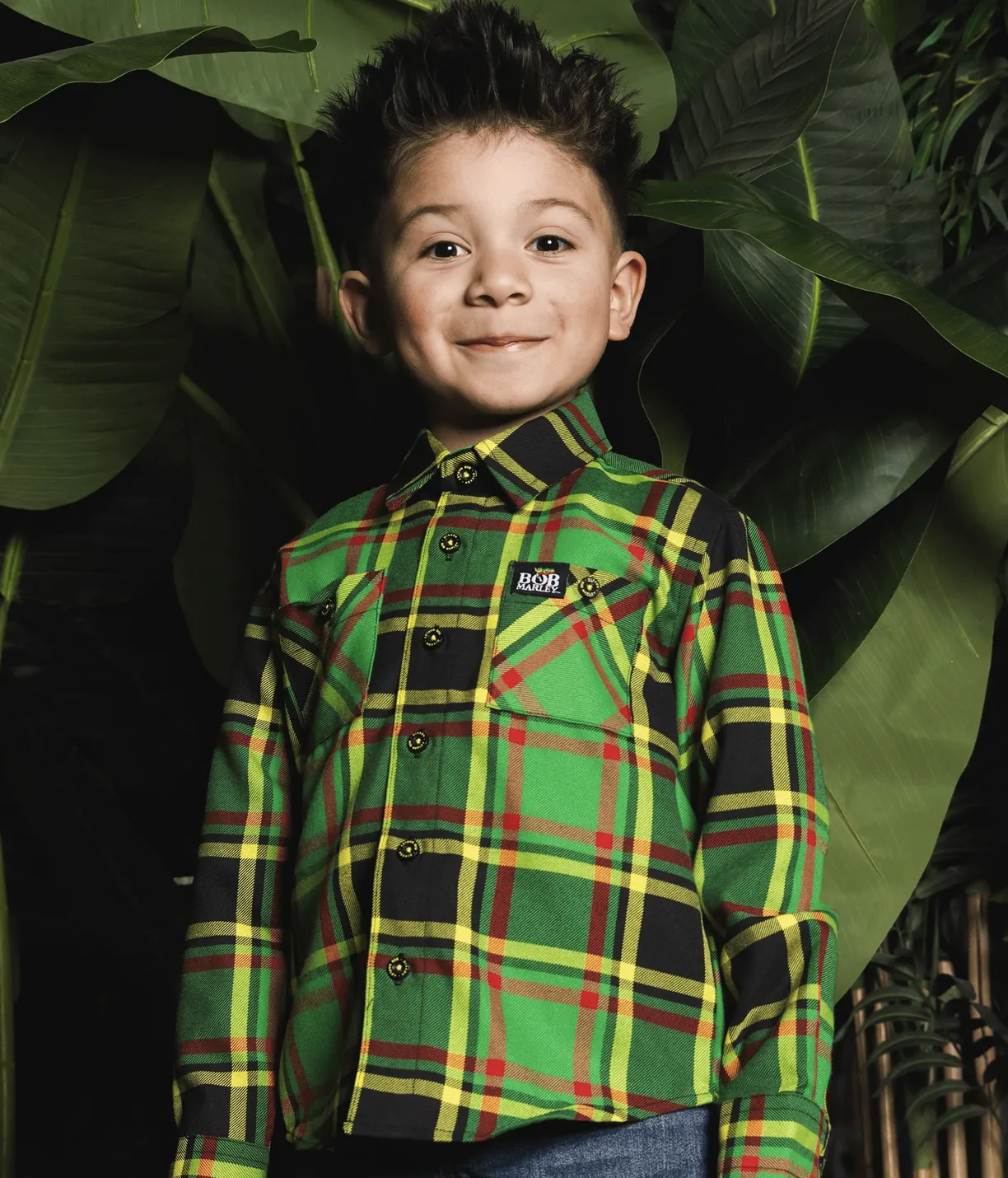 A young boy standing in front of large green tropical leaves, wearing a vibrant green, yellow, and red plaid Bob Marley x Dixxon collaboration flannel with custom branded buttons.