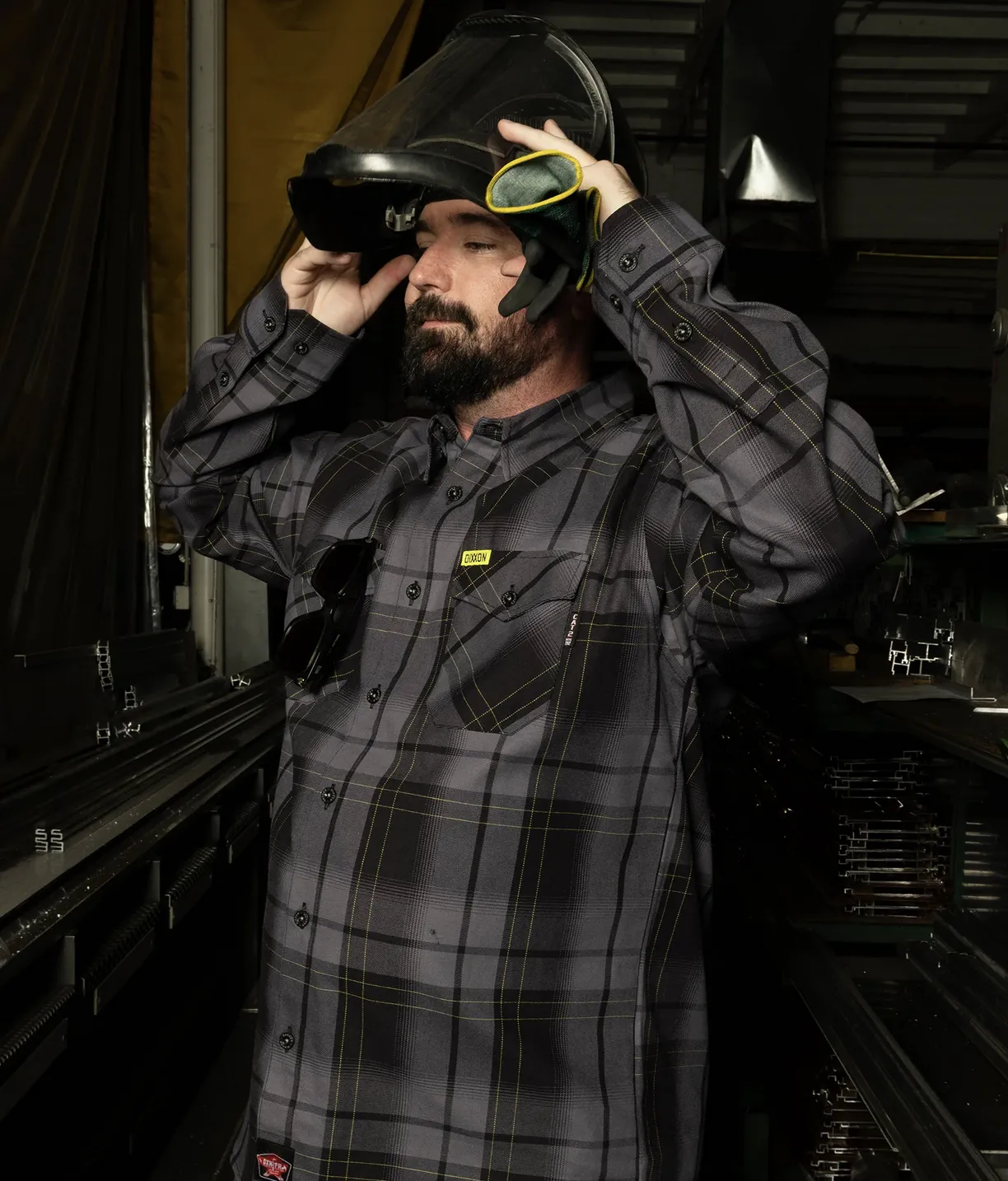 A bearded man in a workshop wears a gray and black plaid Dixxon flannel with yellow accents while adjusting a welding helmet.
