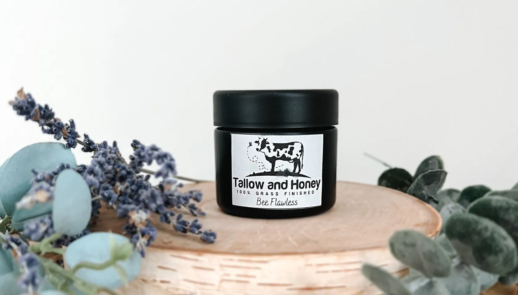 A black jar of "Bee Flawless" 100% grass-finished tallow skincare by Tallow and Honey sits on a wooden pedestal, surrounded by dried lavender to highlight the natural benefits of tallow.