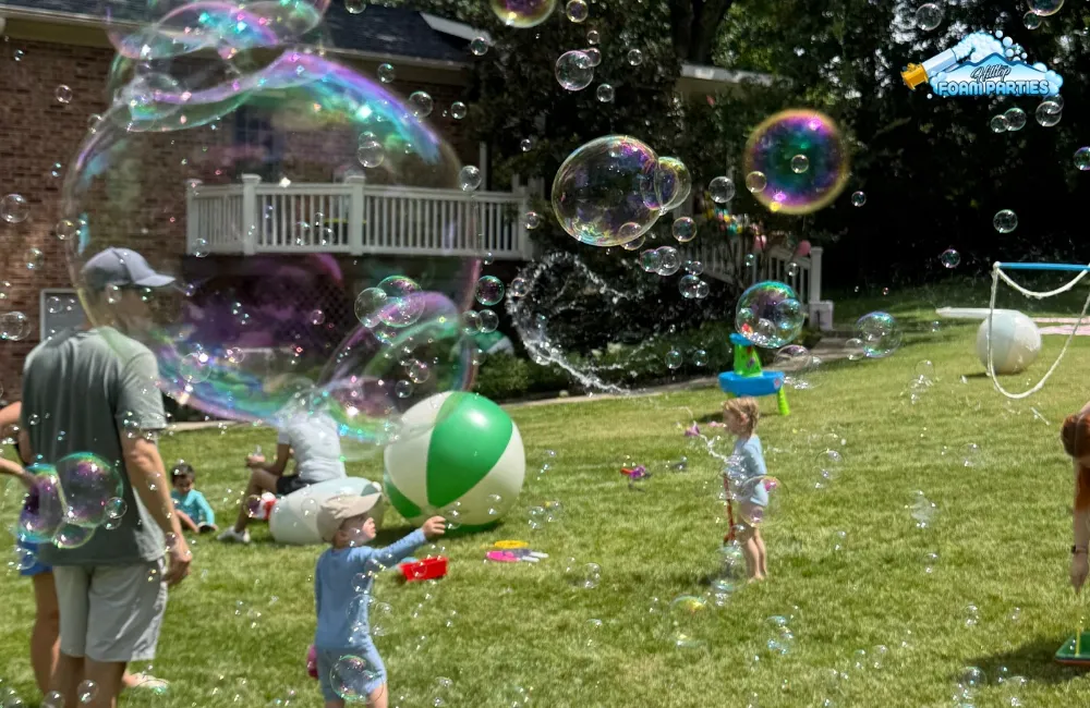 Guests, including children and adults, enjoy giant, colorful iridescent bubbles floating across a sunny residential lawn during a lively outdoor bubble birthday parties event.