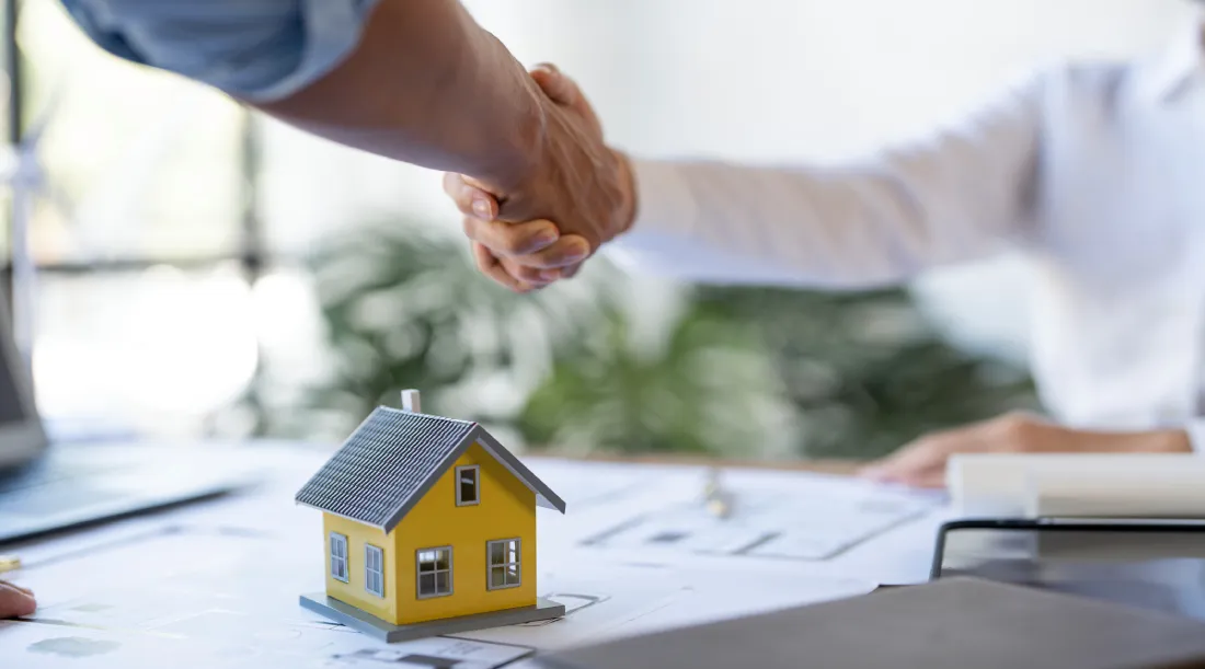 A close-up of a real estate agent and client shaking hands over architectural plans and a yellow house model, symbolizing a successful closing on luxury real estate in Spring Hill and the purchase of new luxury homes.