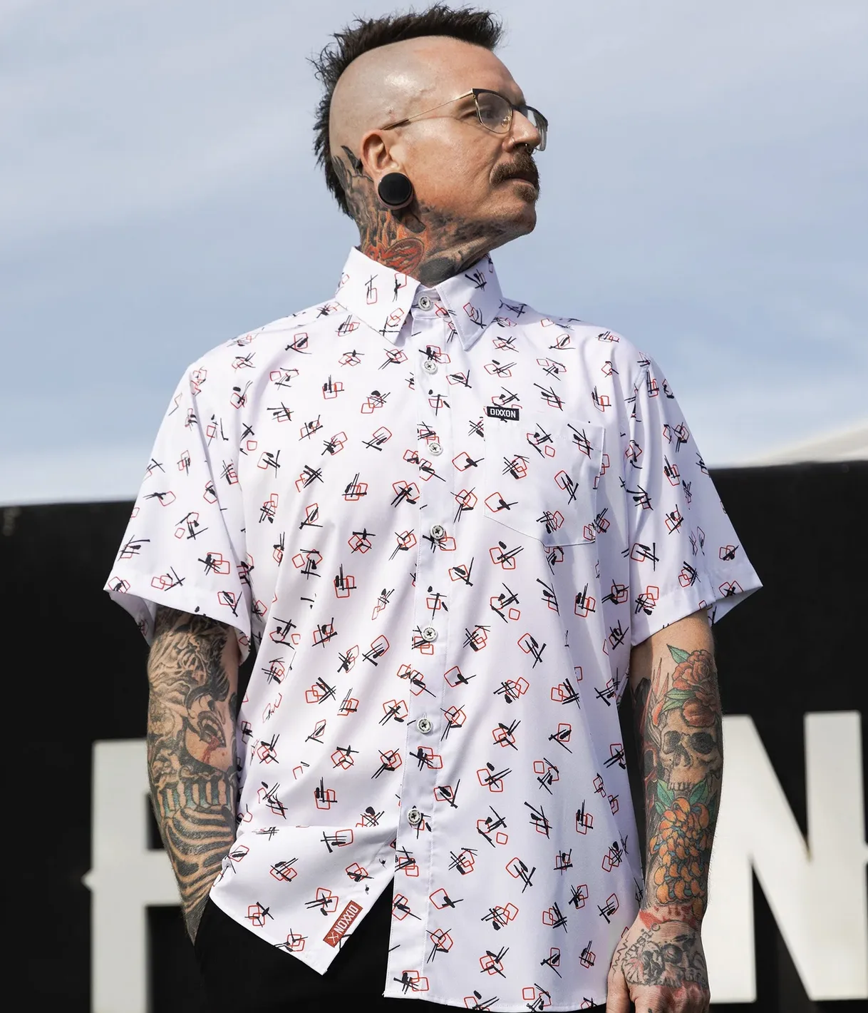 A man with a mohawk and tattoos wears a white Dixxon Party Shirt featuring an edgy, high-contrast geometric pattern in black and red, showcasing the brand’s connection to punk and counter-culture aesthetics.