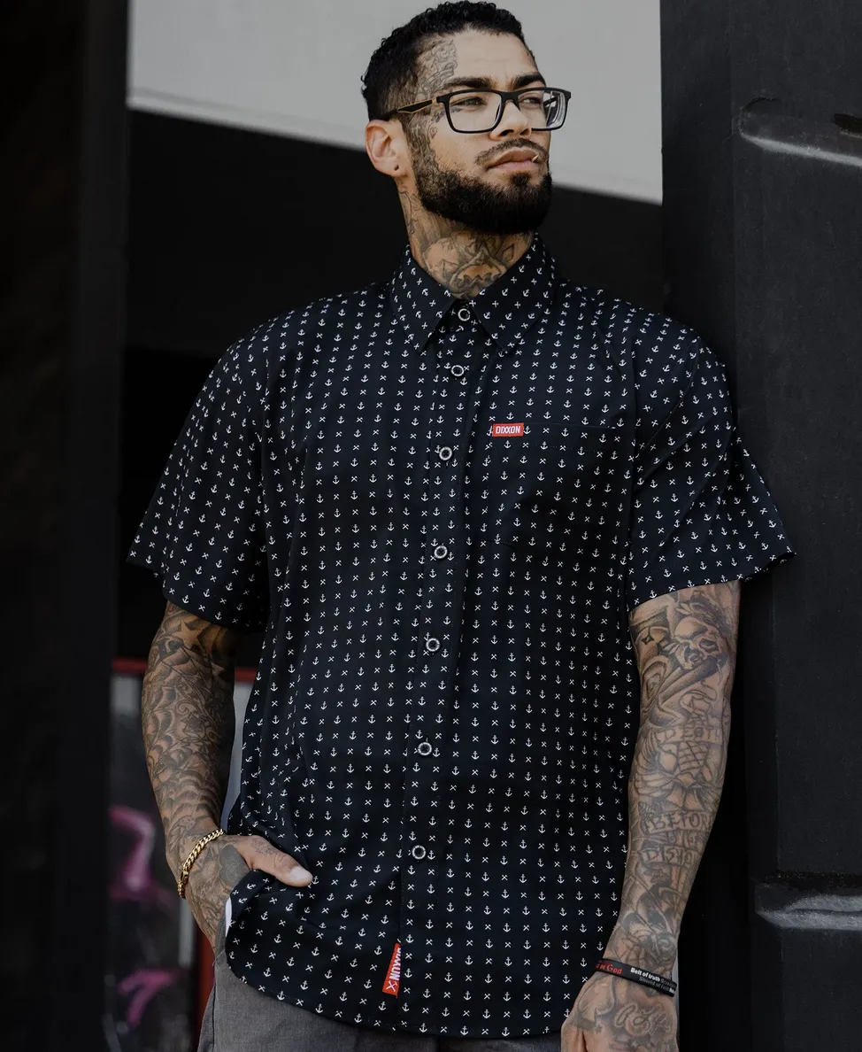 A man with glasses and arm tattoos leans against a dark pillar wearing a black button-down shirt with a repeating white anchor pattern and a red Dixxon logo tag on the pocket.