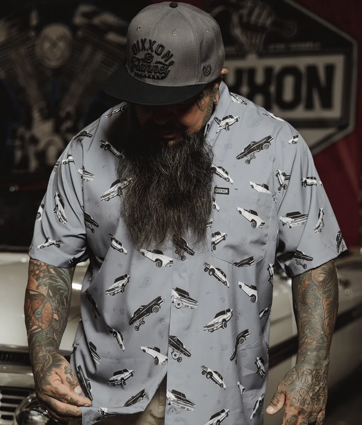 A man with a long beard and extensive arm tattoos wears a grey Dixxon party shirt featuring an all-over pattern of classic black-and-white custom cars. He is wearing a grey-and-black Dixxon "Flannel Company" hat and standing in a garage environment.