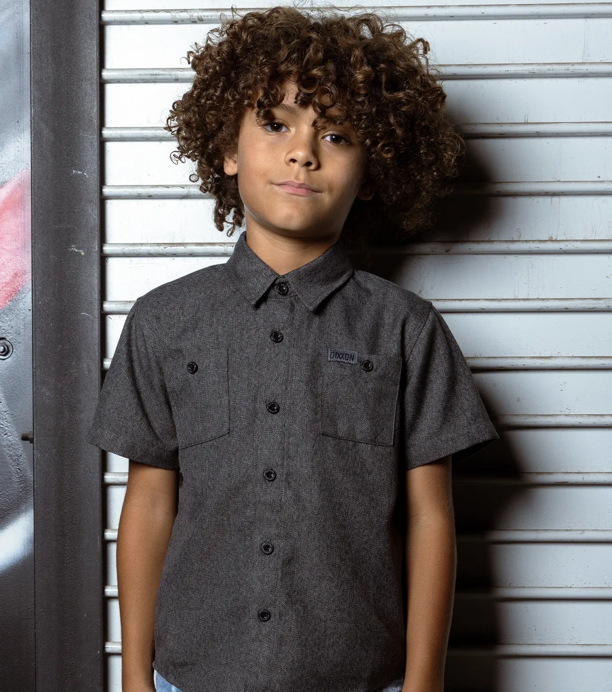 A young boy stands against a rugged metal garage door wearing a clean, charcoal grey Dixxon short sleeve button-up shirt featuring a subtle logo patch.