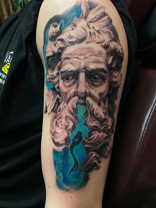 A detailed black and grey portrait of Neptune on an upper arm, with a small silhouette of a mermaid swimming through a vibrant blue ocean scene in his beard.