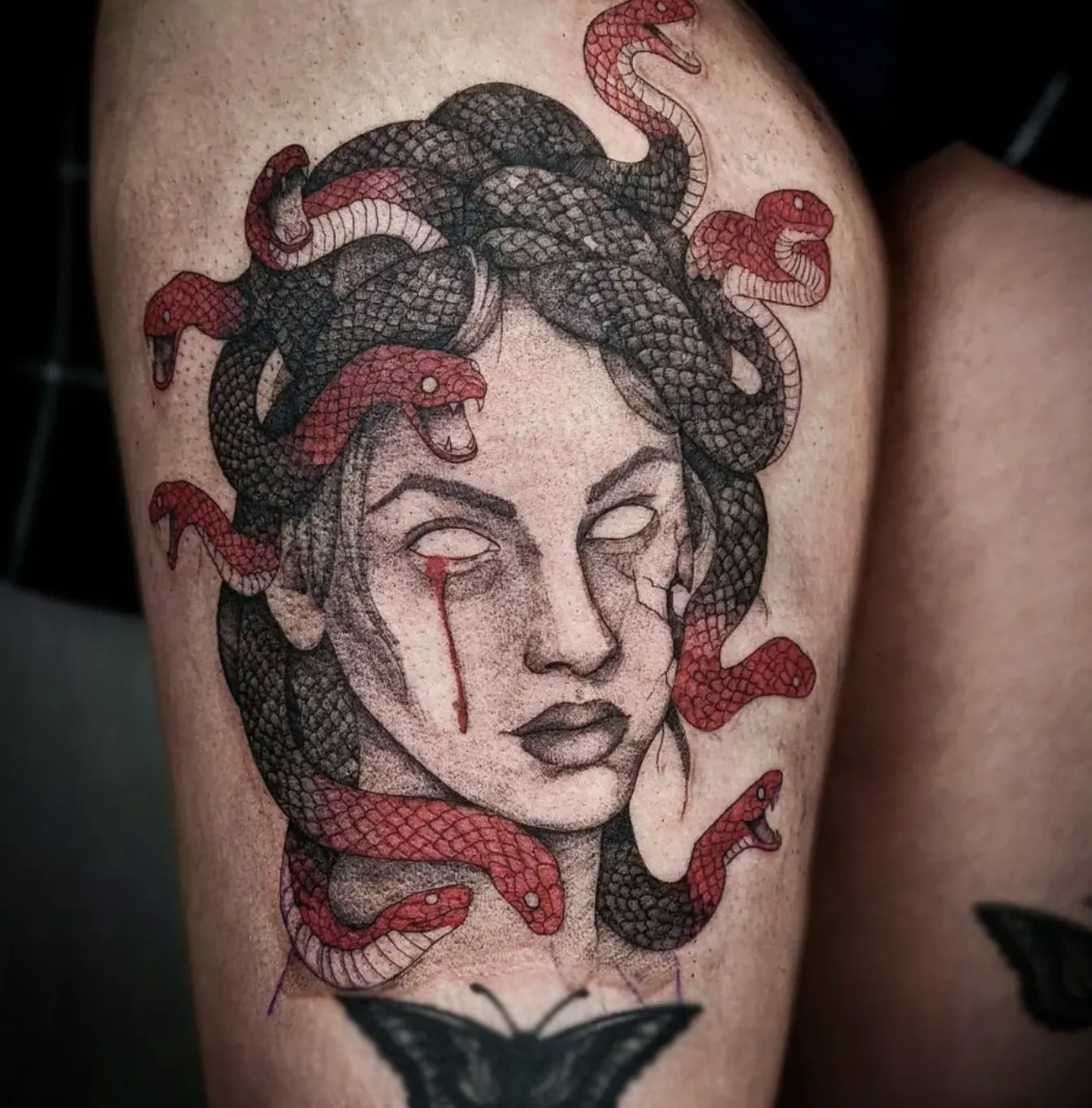 Detailed black-and-grey illustrative tattoo of Medusa on a thigh, featuring a woman with pupilless eyes, a single red tear, and a crown of intertwining black and red snakes.