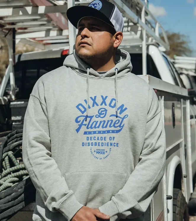 A man wearing a grey Dixxon pullover hoodie with a vibrant blue "Decade of Disobedience" graphic and matching blue drawstring aglets. He is standing in front of a work truck, emphasizing the brand's "Work Hard/Stay Humble" ethos and the apparel's reliability on the job site.