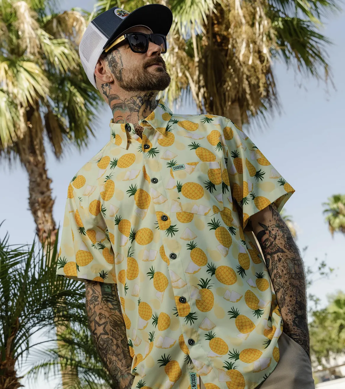 A man with neck and arm tattoos looks off to the side, wearing a pale yellow Dixxon party shirt with a bold, all-over pineapple print. He is standing outdoors in front of tall palm trees under a clear blue sky.