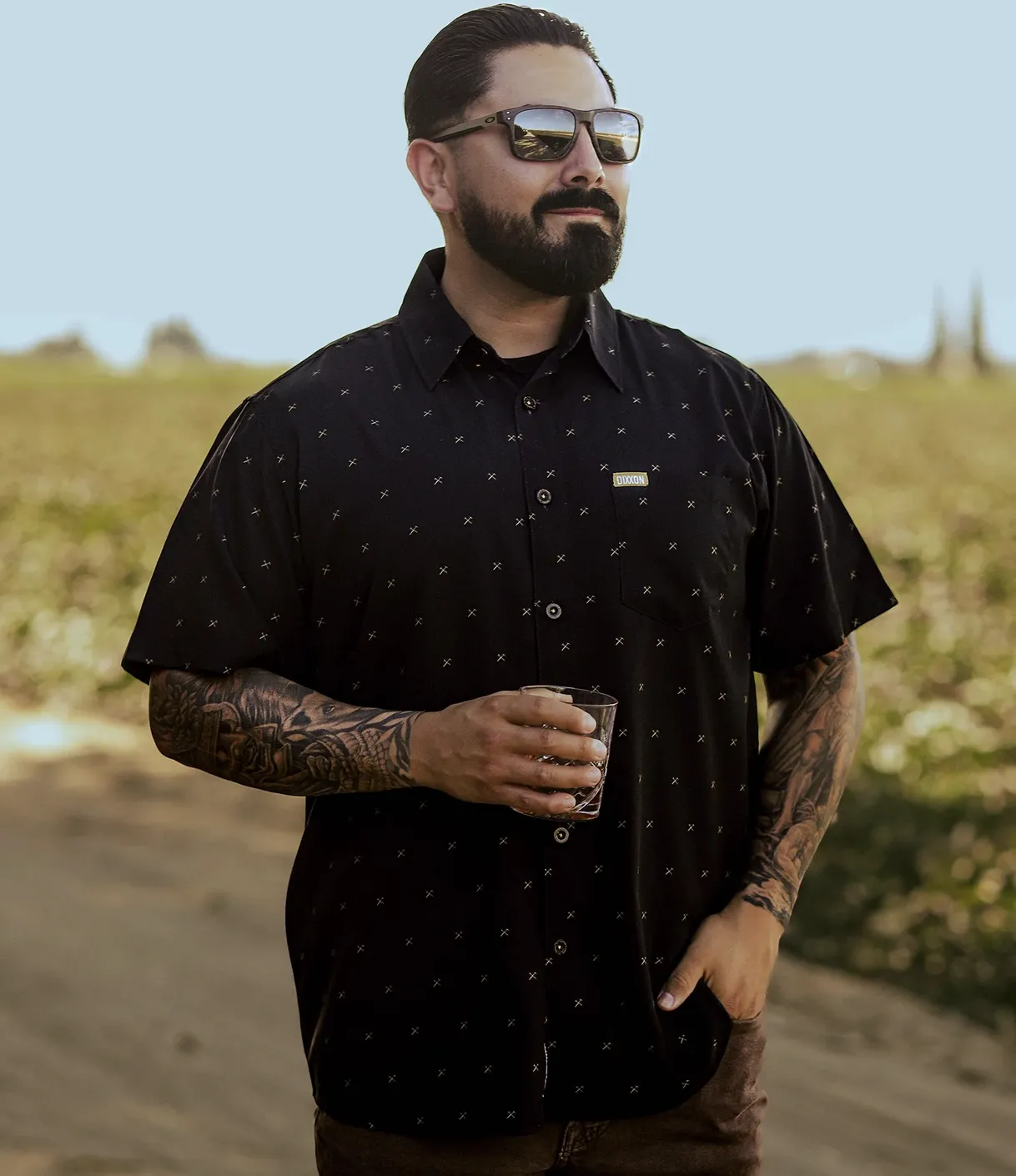 A bearded man in sunglasses stands in a sunlit field wearing a black Dixxon short-sleeve button-up with a subtle, gold-tone repeating cross-tool pattern and a signature brand tag on the chest pocket.