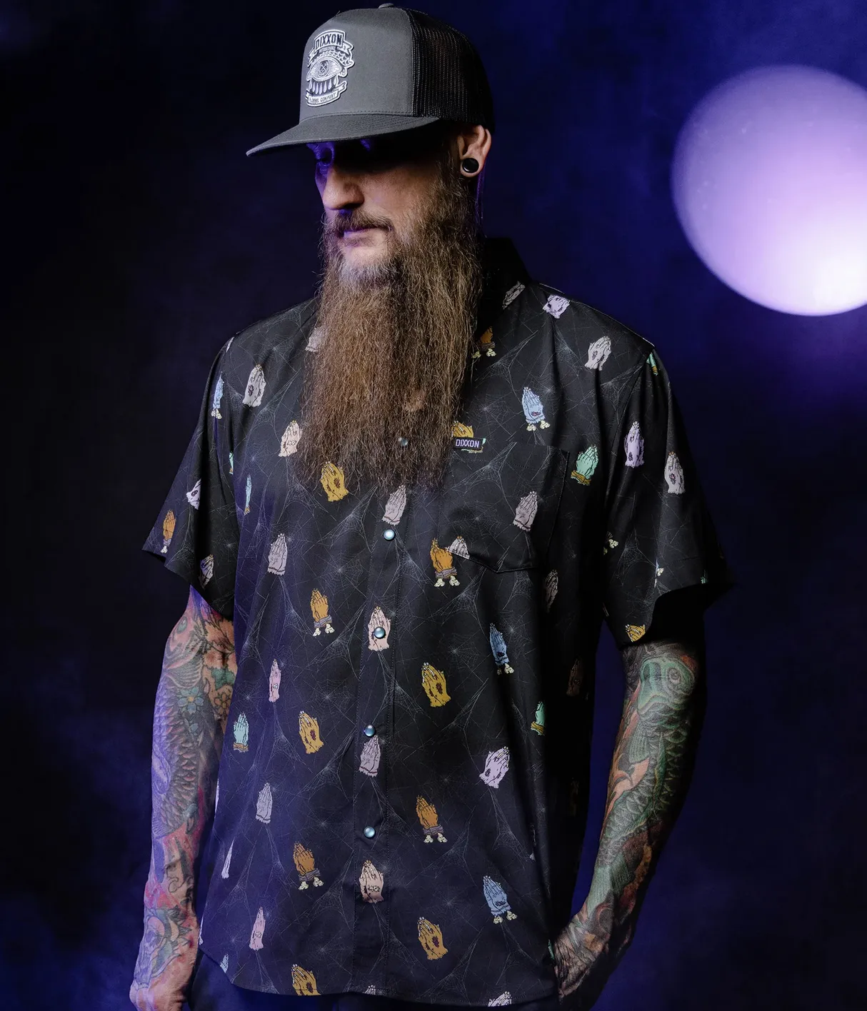 A man with a long beard and tattoos wears a black Dixxon short-sleeve shirt featuring a colorful repeating "praying hands" graphic set against a subtle spiderweb background, paired with a Dixxon branded trucker hat.