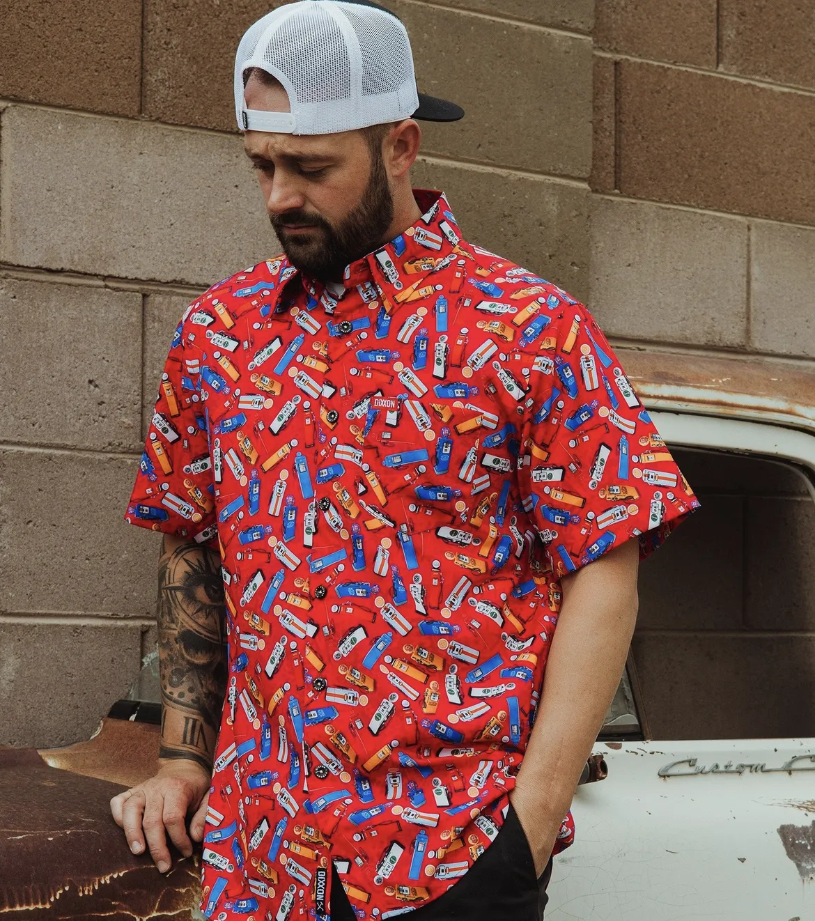 A man with a beard and arm tattoos wears a red Dixxon party shirt featuring a vibrant vintage gas pump and classic car print. He is leaning against a weathered white truck in a casual, straightforward pose.
