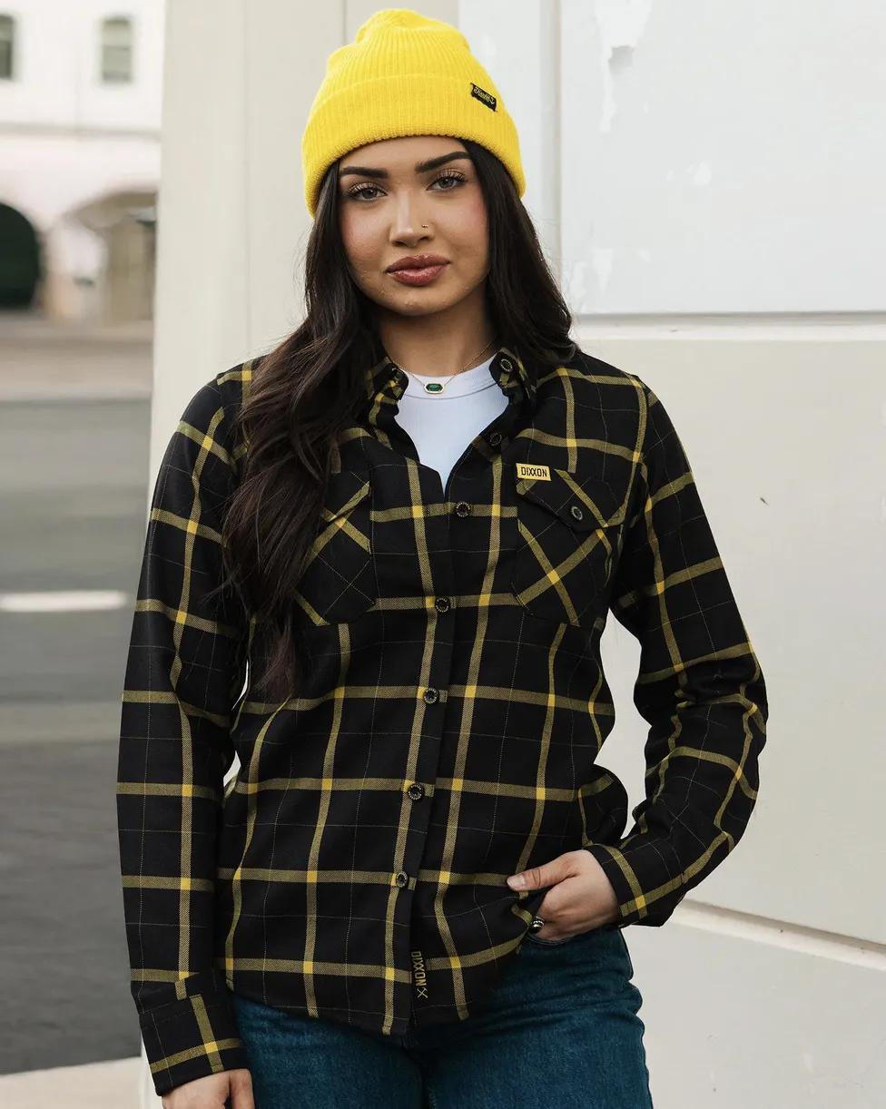 A woman wearing a black flannel with a bold yellow windowpane pattern, paired with a bright yellow beanie. The shirt features a yellow Dixxon brand tag on the chest pocket. She is posed against a clean, white urban background.