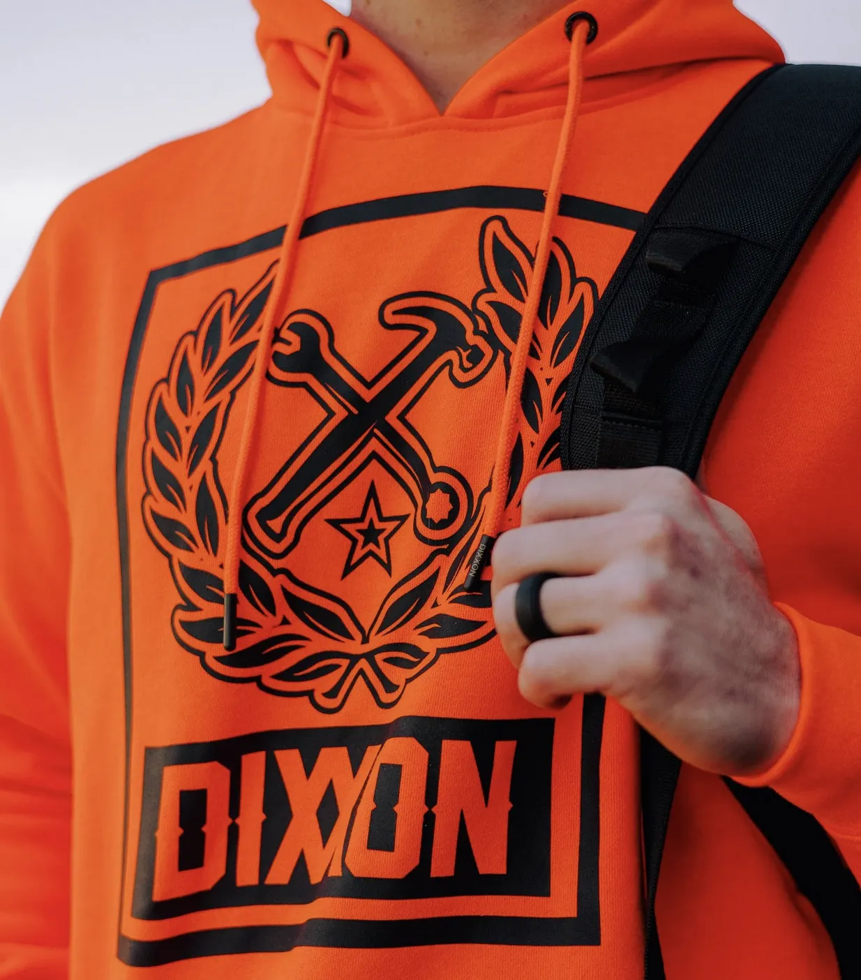 Close-up of a bright orange pullover hoodie featuring a large black "Box Logo" with the Dixxon name and a crest containing a crossed wrench and hammer.