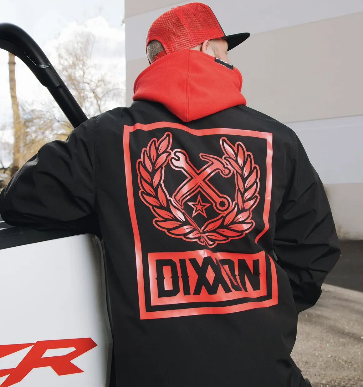 A person viewed from the back, leaning against the side of a white vehicle while wearing a red hoodie and a black jacket with a large red graphic on the back. The graphic features a star and crossed tools encircled by a wreath, with the "DIXXON" brand name in a bold, stylized font below it.
