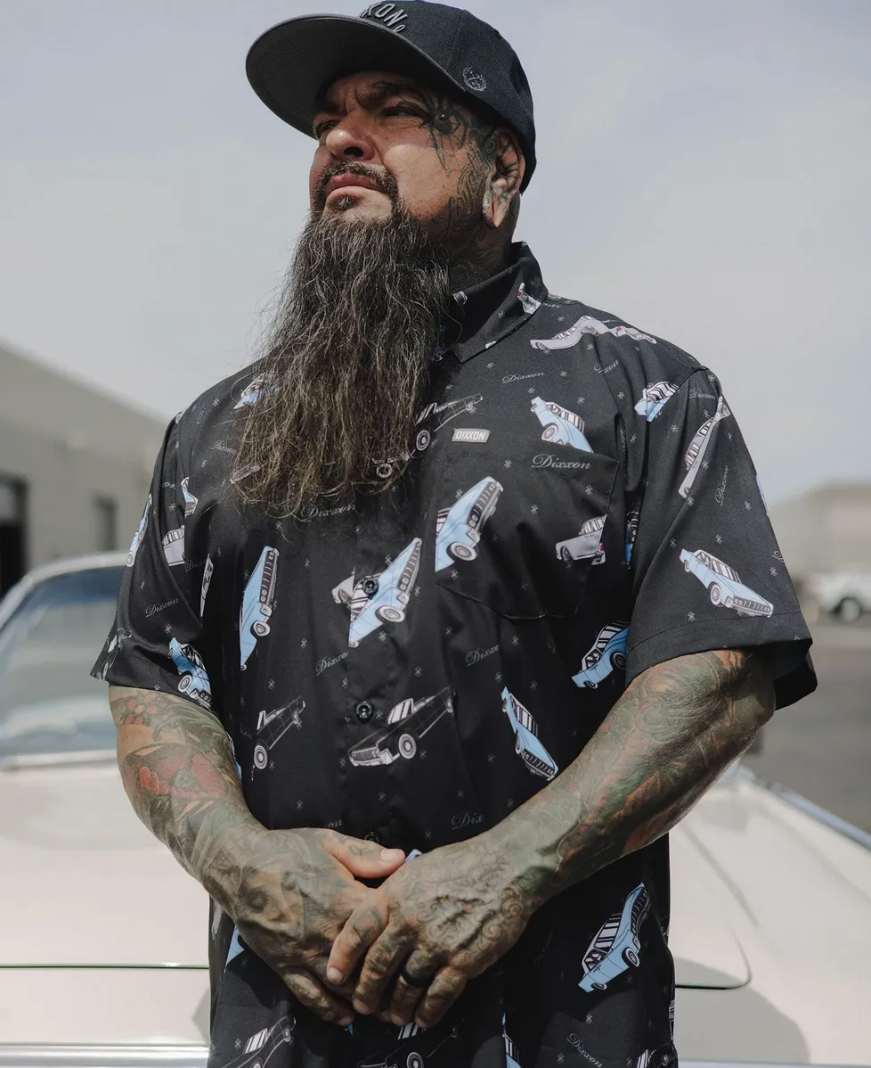 A man with a long beard and extensive arm tattoos wears a black party shirt featuring a repeating lowrider car print. He stands confidently in front of a classic car, looking towards the horizon.