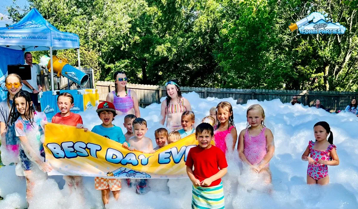 A large group of happy children posing together in a thick layer of bubbles during a summer foam event. Two children in the front hold a yellow banner that reads "best day ever," while a professional foam cannon and branding for hilltop foam parties are visible in the background against a line of green trees.