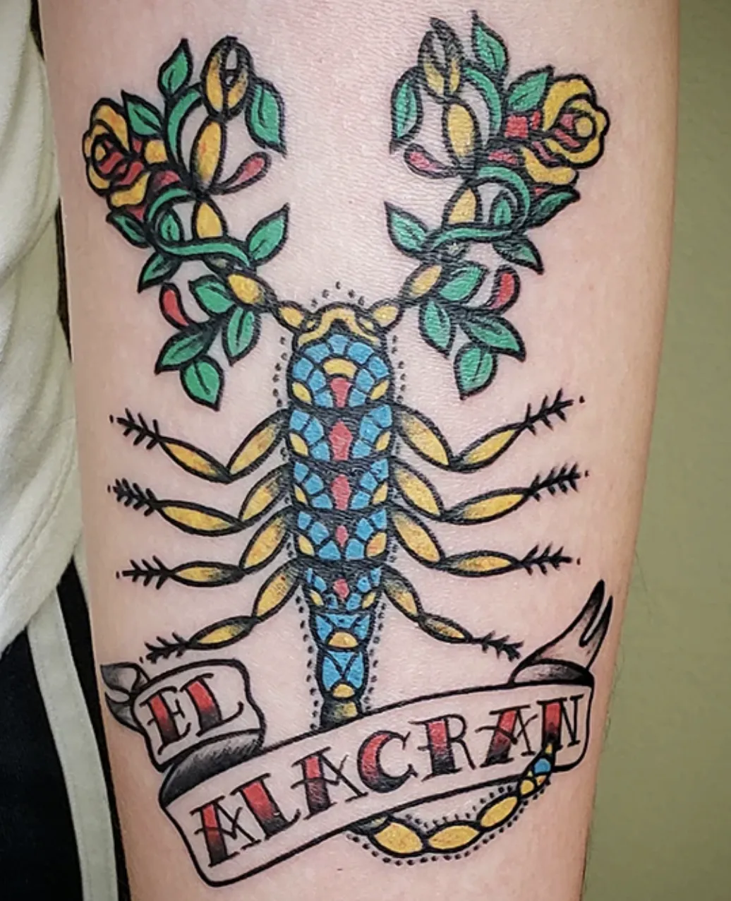 Bold American traditional style tattoo on an arm of a scorpion (labeled "EL ALACRAN"), where the claws are replaced by blooming yellow roses and green leaves.