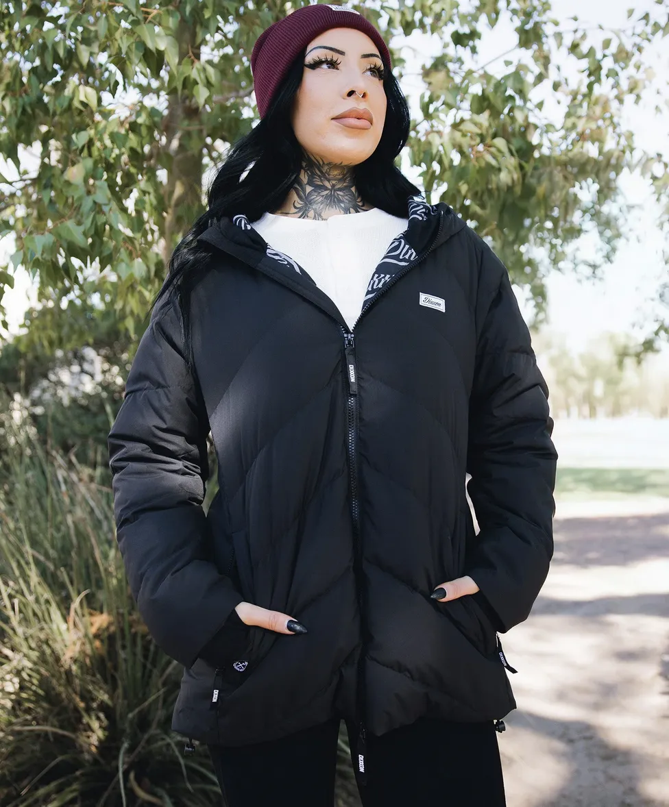 A woman wearing a burgundy beanie and a long black quilted Dixxon parka with a hood and white Dixxon logo patch. She has visible neck tattoos and is posing outdoors in a wooded area.