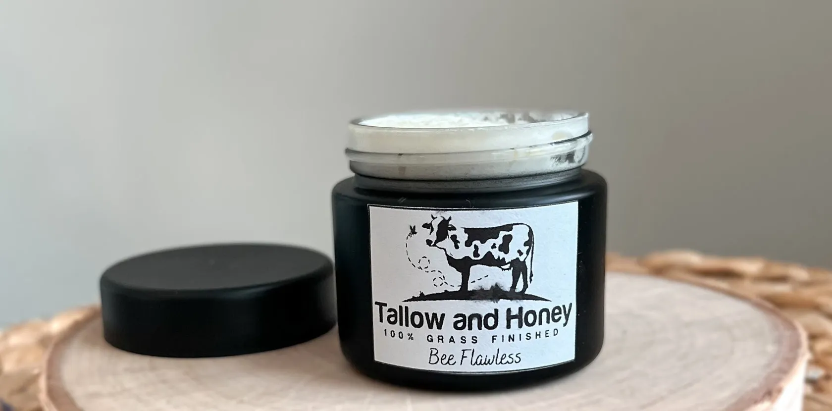 An eye-level view of a black glass jar labeled "Tallow and Honey Bee Flawless," containing 100% grass-finished tallow cream and displayed on a light wooden coaster.