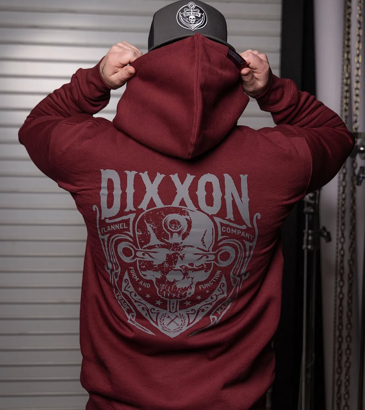 The back view of a heavyweight burgundy Dixxon hoodie featuring a large grey "DIXXON FLANNEL COMPANY" skull and anchor graphic with the "Form and Function" slogan. A man is shown pulling up the hood, showcasing the durable fabric and bold counter-culture aesthetic.