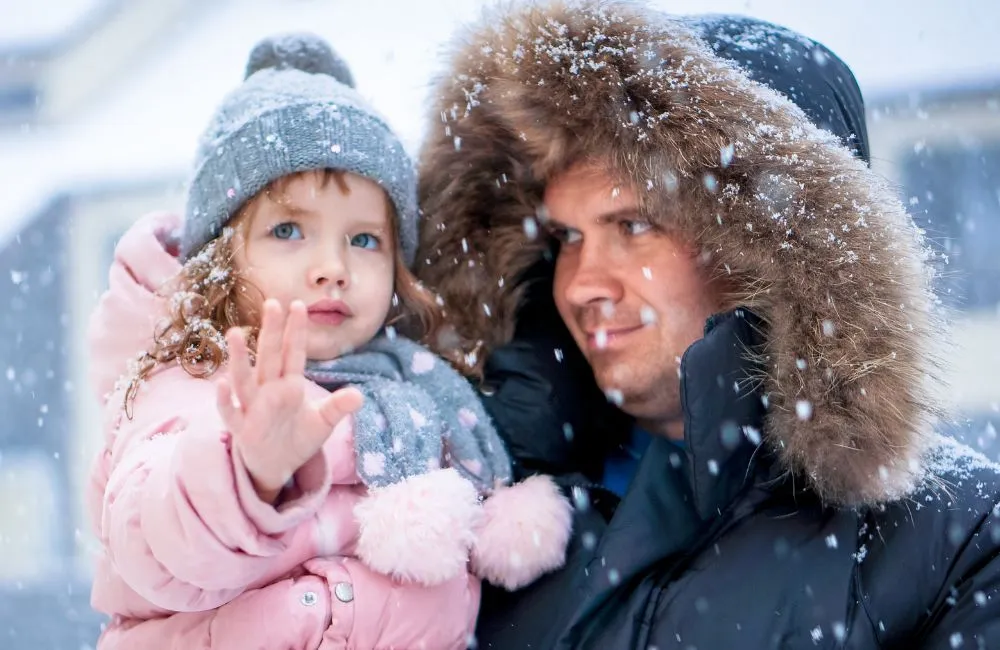 A close-up image of a father holding a young child outside in the snow. The father wears a black winter coat with a fur-lined hood, and the child wears a pink puffer coat, a gray pom-pom knit hat, and a polka-dotted scarf and gloves. The child, with reddish-brown curly hair and blue eyes, is looking up and raising a hand, seemingly trying to catch the snowflakes. The snow appears to be falling heavily, possibly from natural snowfall or a professional setup using snow machine rentals.