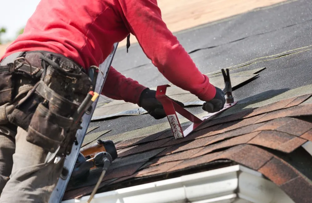 A roofer wearing a red shirt, grey pants, and a tool belt is kneeling on a partially shingled roof, securing a temporary safety bracket (a roof jack) into the exposed tar paper and partially installed asphalt shingles, illustrating the work involved with various roofing choices.