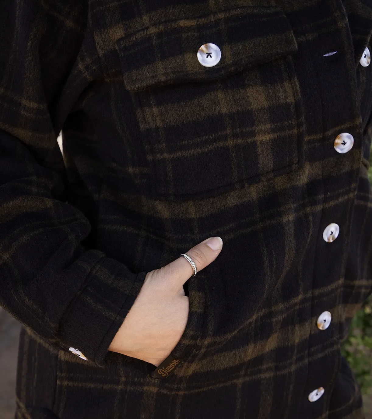 A close-up view of a heavy, olive green and black plaid fabric featuring a button-down front with large, marbled white buttons. A hand with a silver ring on the thumb is resting in a side pocket.