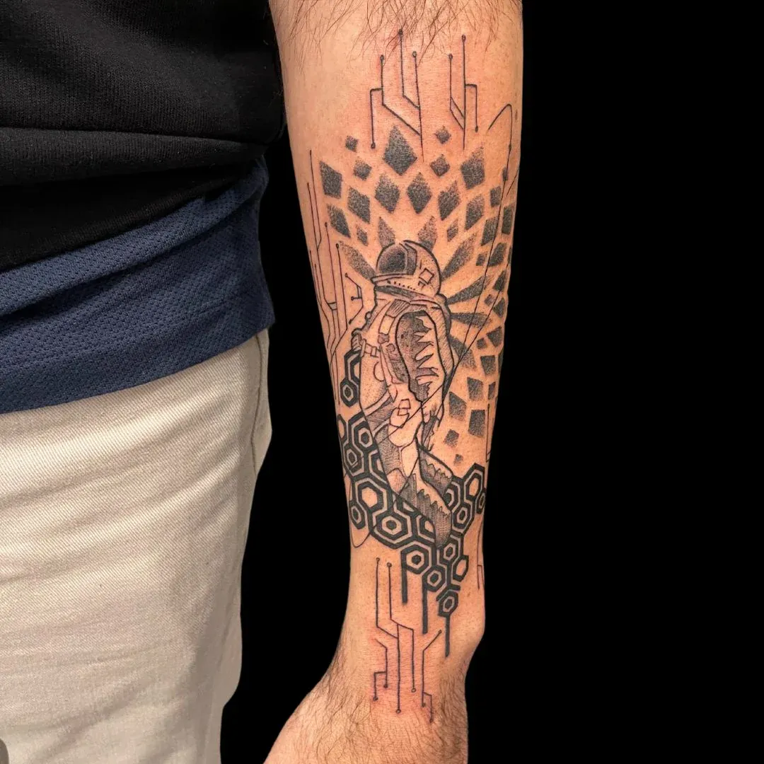 A stylized black ink tattoo of an astronaut on a forearm, integrated into a complex background of geometric diamond patterns and motherboard-like circuitry.