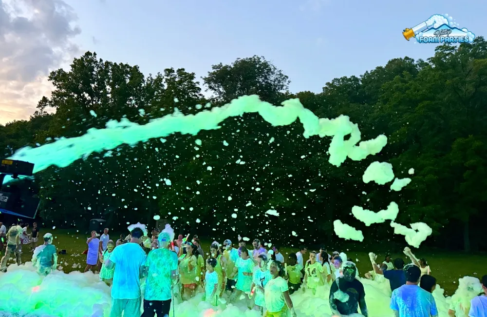 A large group of people enjoying an outdoor foam glow party in a grassy area surrounded by trees at dusk, as a machine shoots a stream of glowing green foam over the crowd.