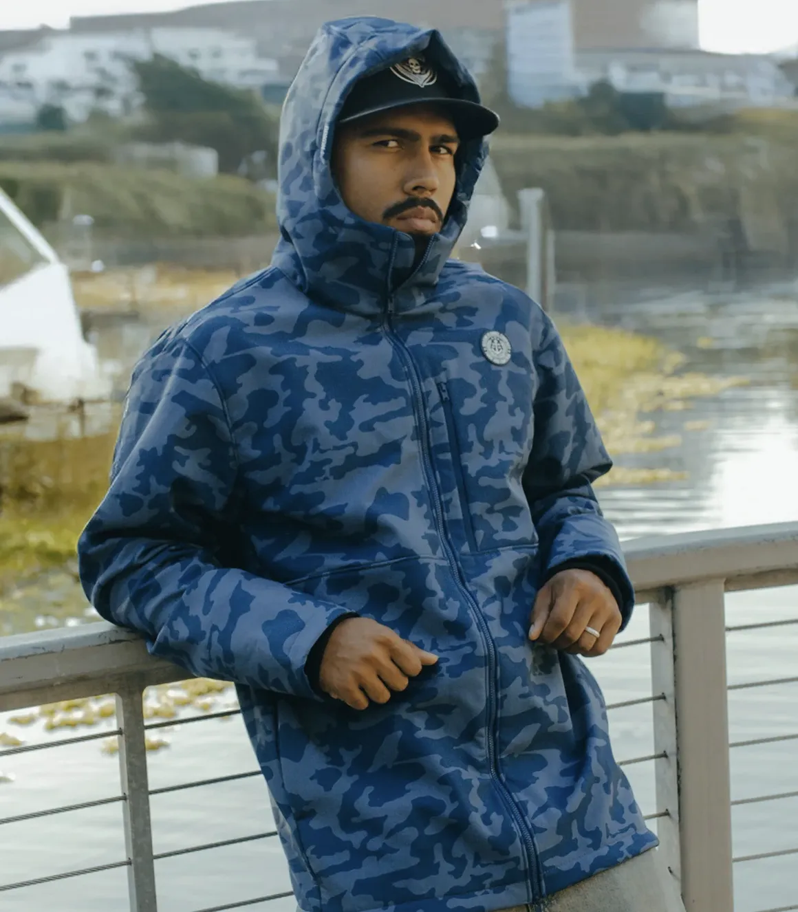 A person leaning against a railing by the water wearing a blue camouflage-patterned Anchor Jacket with the hood up.