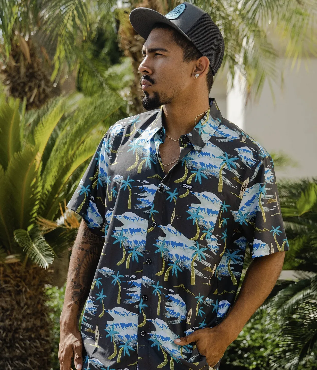 A man wears a dark-colored, short-sleeved Dixxon party shirt featuring an "easy fluid fabric" with a vibrant tropical print of palm trees, waves, and surfboards. He is also wearing a charcoal Dixxon trucker hat and standing in front of palm trees.