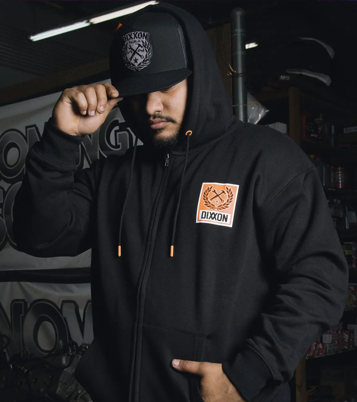 A close-up of a man in a black Dixxon full-zip hoodie with custom orange eyelets and aglets, paired with a matching black and grey Dixxon patch hat. He is adjusting his hat in a workshop setting, highlighting the gear's rugged functionality and style.