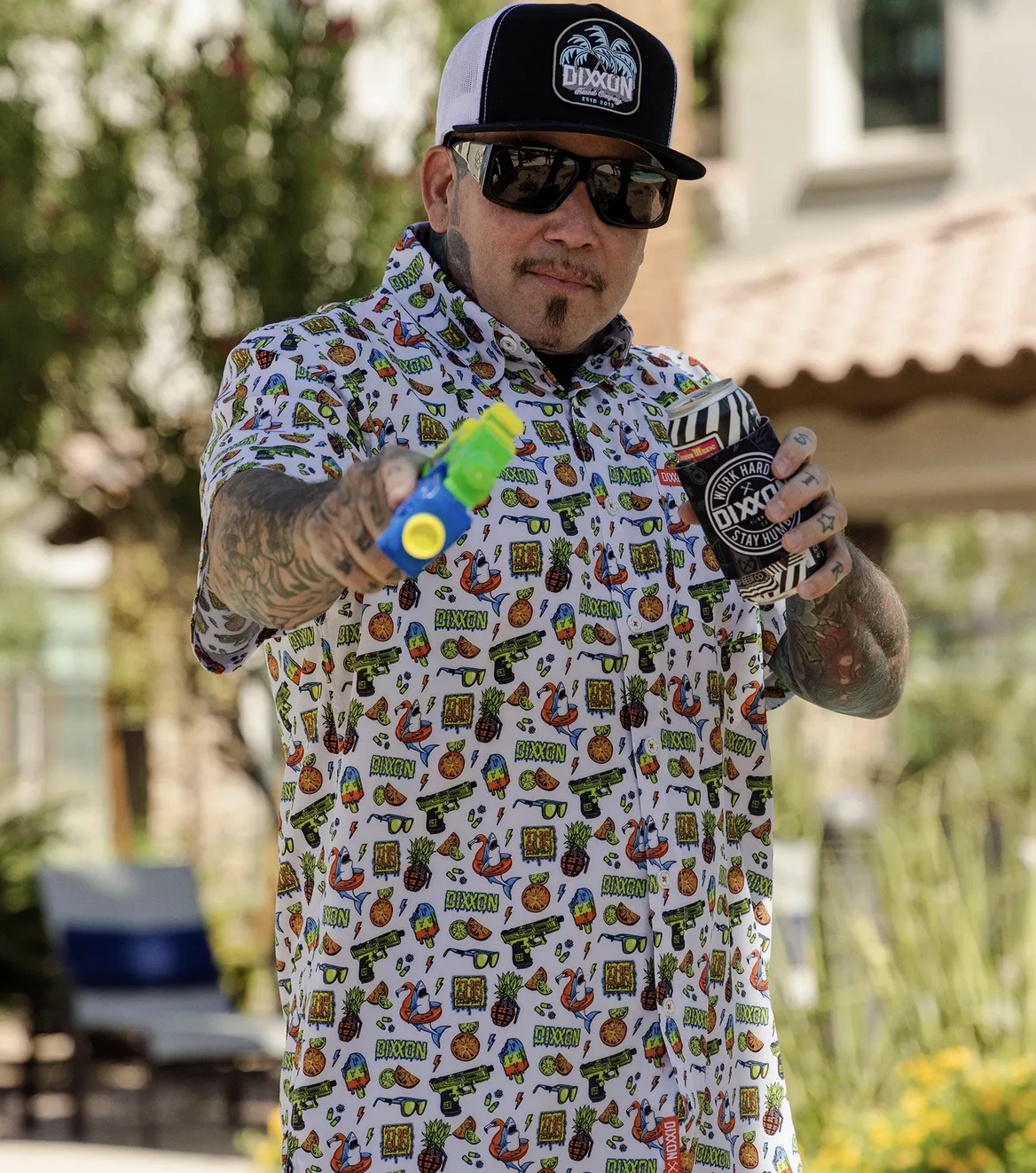 A man holding a water gun and a drink in a Dixxon koozie wears a white party shirt covered in playful summer graphics like pineapples, sharks, and sunglasses. He is wearing a black and white trucker hat with a palm tree logo.