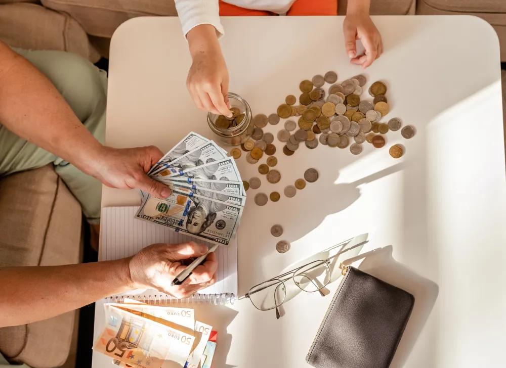 An overhead view of two pairs of hands—one holding US $100 bills and writing, the other placing coins into a jar—while counting Personal Finance money on a white table, possibly saving for a future Roof Repair project.