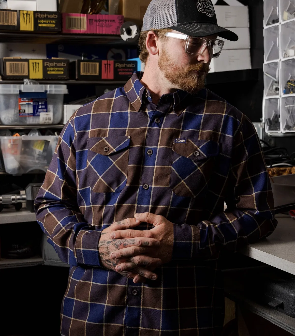 A man in a workshop setting with tattooed hands, wearing a navy blue and chocolate brown plaid Dixxon flannel designed for durability and a hard-working lifestyle.