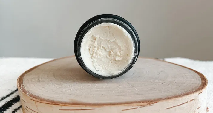 A top-down view of a black glass jar filled with creamy white Tallow Products, displayed on a rustic wooden slice against a neutral background.