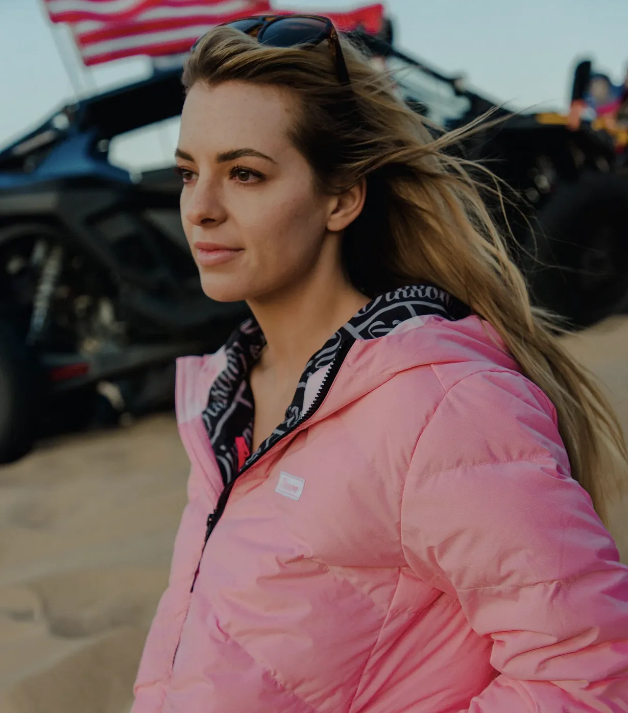 A woman with blonde hair wearing a bright pink Dixxon Ridge Down jacket outdoors with a vehicle and American flag in the background. The jacket includes a protective zipper garage at the neck and a custom Dixxon logo patch on the left chest, showcasing a blend of form and high-performance function for dynamic adventures.